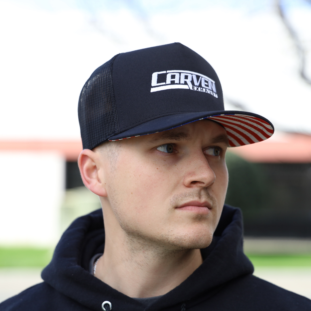 A man looking to the side while wearing a black hat with a white Carven Exhaust logo and an American flag brim